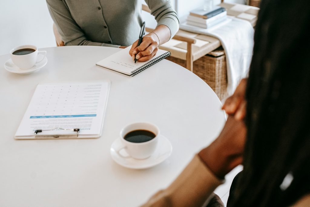 Réunion professionnelle autour d'une table blanche où deux personnes discutent d'une stratégie e-commerce, l'une prenant des notes dans un carnet pendant que des documents marketing et deux tasses de café sont posés sur la table, représentant la planification d'un projet de vente en ligne pour artisans.