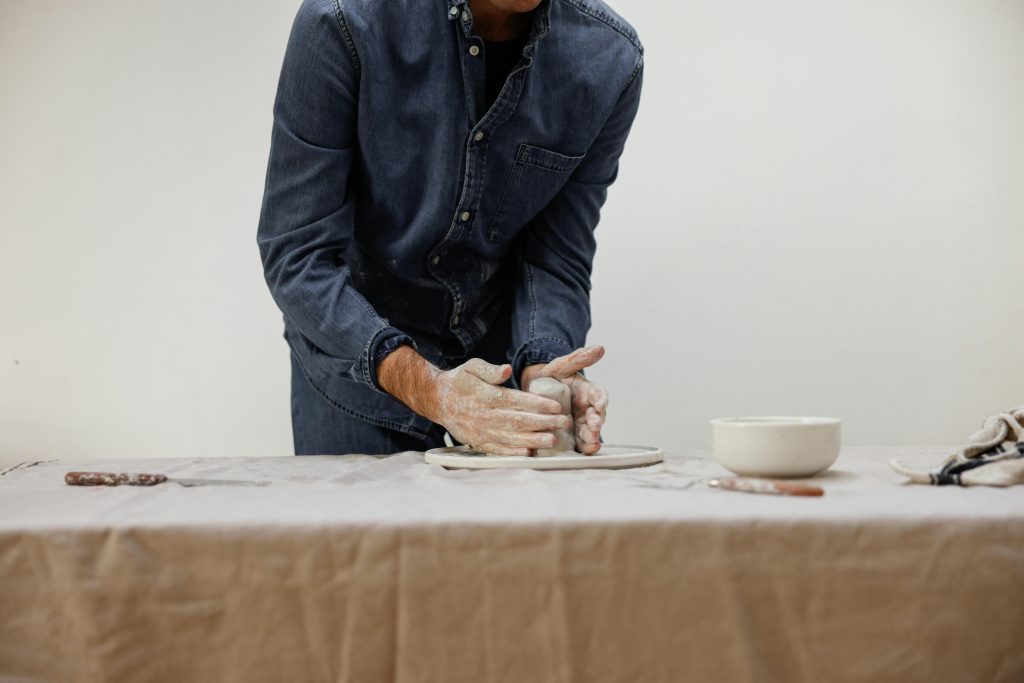 Artisan céramiste en chemise denim façonnant l'argile sur un tour de potier, mains expertes et couvertes d'argile modelant une pièce sur une table de travail neutre, illustrant le savoir-faire artisanal et la création manuelle pour un site internet d'artisan d'art mettant en valeur les métiers de la céramique et de la poterie.