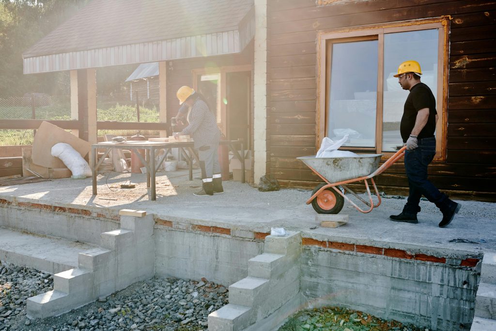 "Chantier de construction en cours où deux artisans du bâtiment travaillent sur une terrasse en béton. À droite, un ouvrier portant un casque jaune de sécurité pousse une brouette avec des matériaux, tandis qu'à gauche, un second artisan travaille sur une table avec des outils. La maison en bois est en phase de rénovation avec une nouvelle fenêtre installée. Cette scène illustre parfaitement le quotidien des professionnels qui pourraient optimiser leur gestion grâce à un logiciel artisan bâtiment permettant le suivi chantier numérique, la gestion administrative des travaux et l'élaboration de devis-facturation précis. Une solution digitale adaptée offrirait un gain de temps considérable pour ces artisans en centralisant toutes les informations du projet.