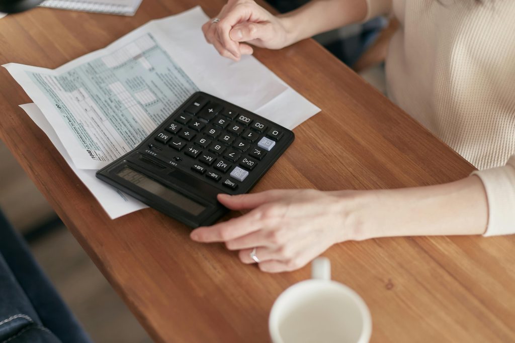 Artisan calculant un devis sur un bureau en bois avec une calculatrice noire posée sur des documents de facturation. Les mains d'une personne manipulent la calculatrice tout en examinant attentivement les papiers, illustrant la gestion administrative quotidienne que peut simplifier un logiciel de devis facturation pour artisans du bâtiment. Une tasse blanche est visible dans le coin inférieur de l'image, évoquant l'environnement de travail d'un entrepreneur ou d'une PME du BTP.