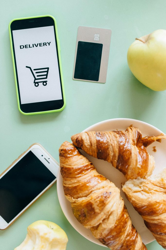 Application de livraison alimentaire affichée sur un smartphone avec icône de panier d'achat, à côté d'une carte bancaire, de croissants frais sur une assiette et de pommes sur une surface vert menthe. Illustration parfaite de la digitalisation du commerce alimentaire permettant la gestion des commandes en ligne, la fidélisation client numérique et la gestion des stocks en temps réel pour les commerces locaux.