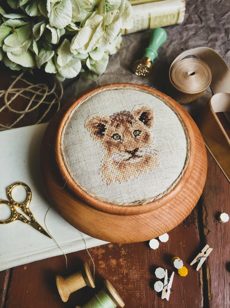 Espace de travail chaleureux d'un artisan brodeur avec un tambour en bois contenant une broderie détaillée d'un lionceau, entouré d'outils de création comme des ciseaux dorés, une bobine de ruban, un sceau à cacheter et des accessoires de couture, idéal pour un site internet d'artisan d'art mettant en valeur le savoir-faire artisanal et la minutie du travail fait main.